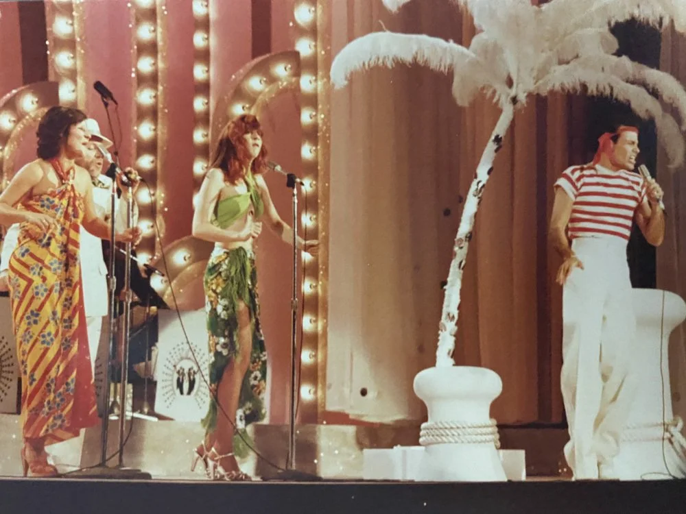 Four performers on a stage during a show, with vintage-style lights, a wooden backdrop, and an artificial palm tree prop.