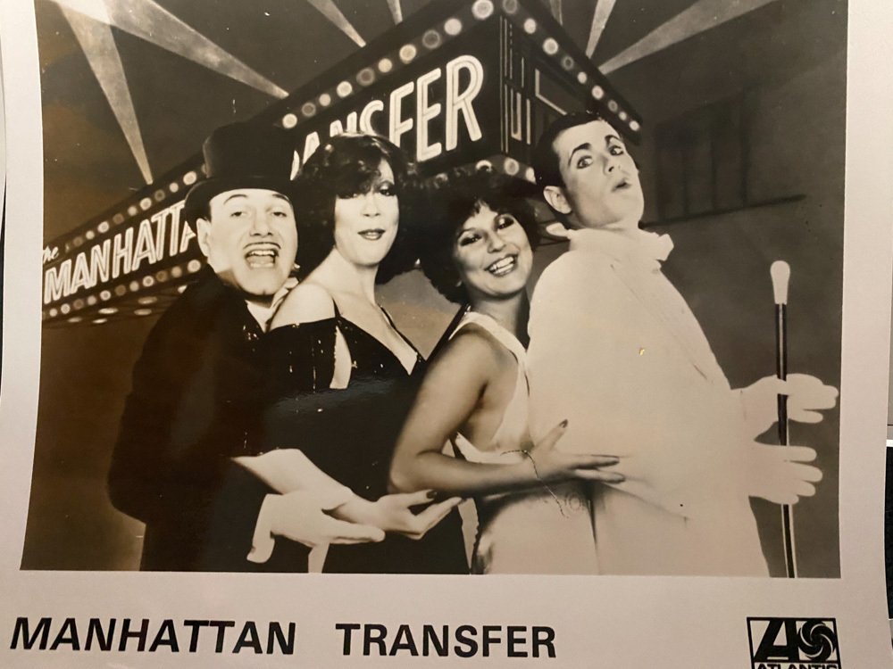 Four performers in vintage costumes standing in front of a Manhattan Transfer marquee, possibly at a theatrical or nightclub setting, in black and white.