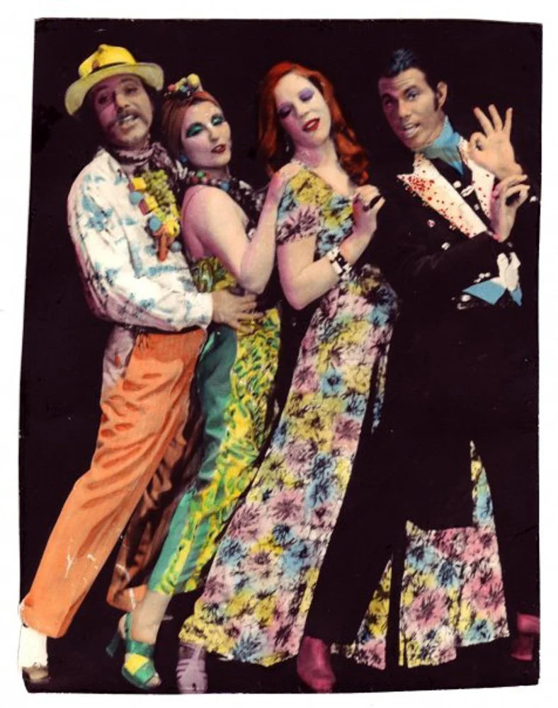 Four people dressed in colorful, vintage-style clothing posing against a black background.
