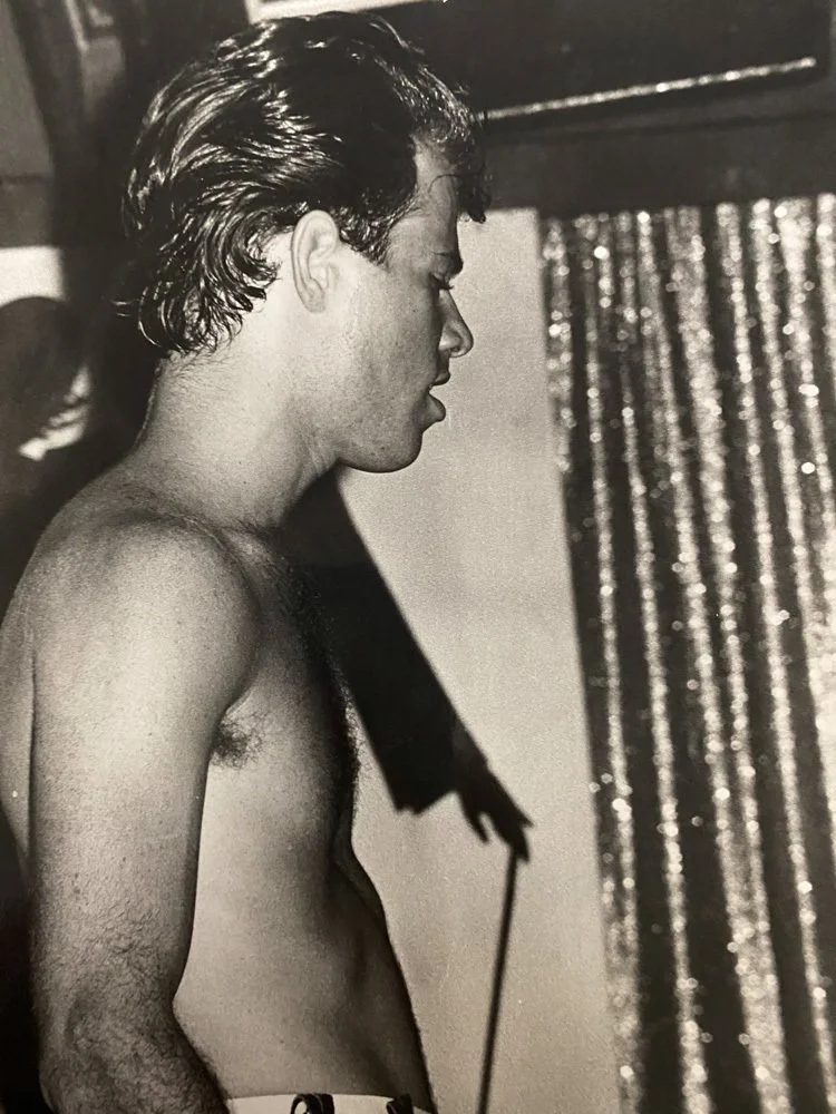 Black and white photograph of a shirtless young man with slicked-back hair, standing in profile.