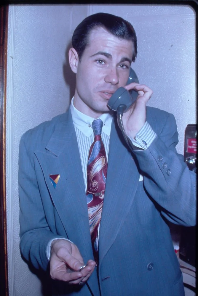A young man in a gray pinstripe suit with a colorful tie, short dark hair, and a brooch on his lapel, speaking on a payphone, with a slightly serious expression.