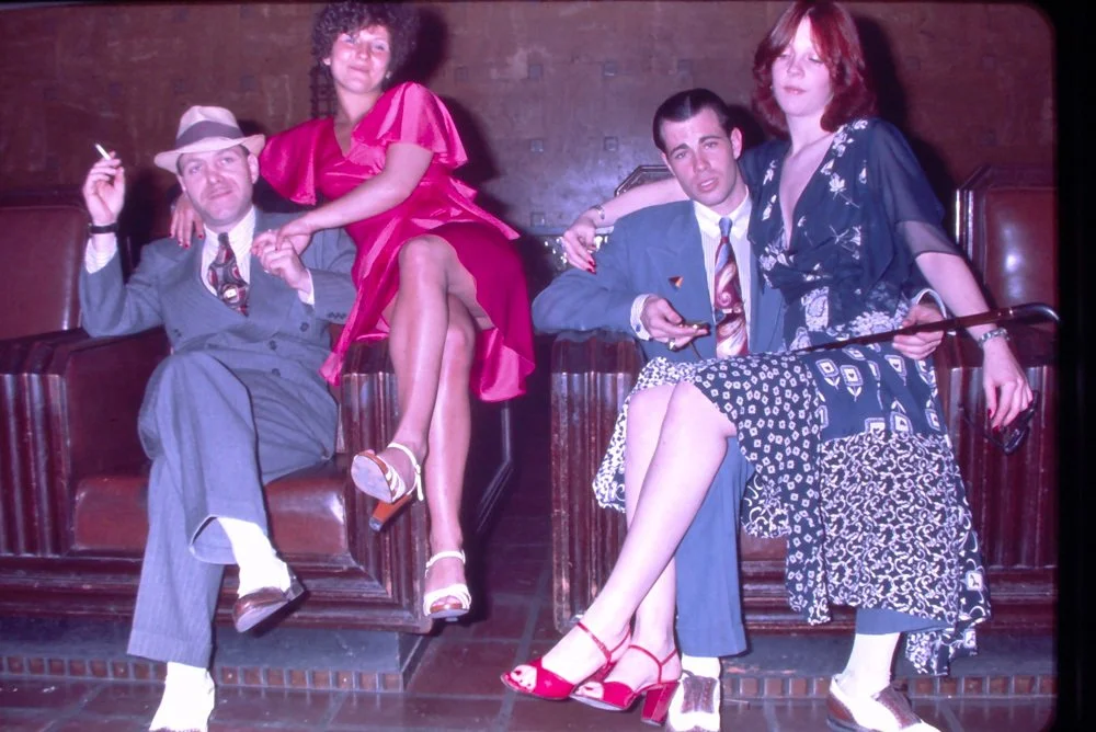 Four young adults with dyed hair and vintage clothing sitting on leather booths, two men and two women, in a dimly lit bar or nightclub with brick walls.