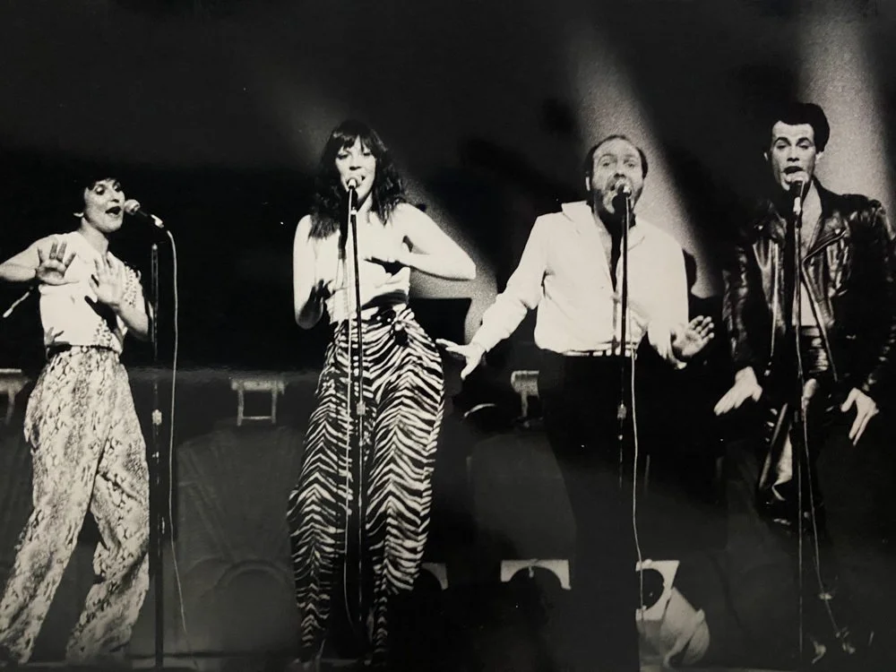 Black and white photo of four performers singing on stage with microphones, two women on the left and two men on the right.