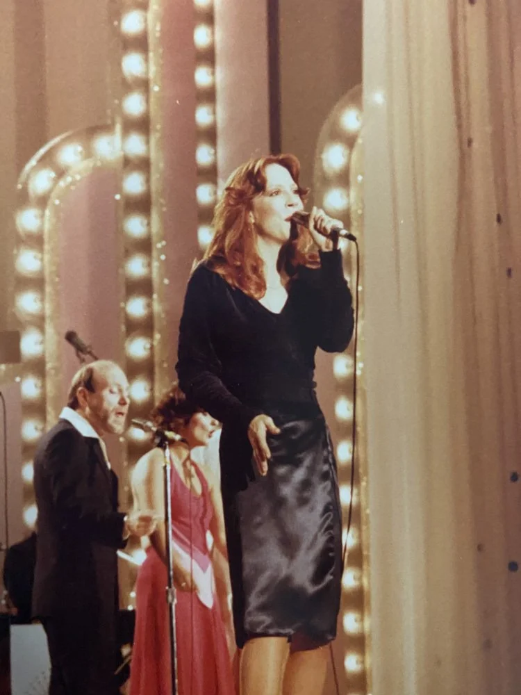 A woman with red hair singing into a microphone on stage with two other people in the background, one man and one woman, near a brightly lit curtain.