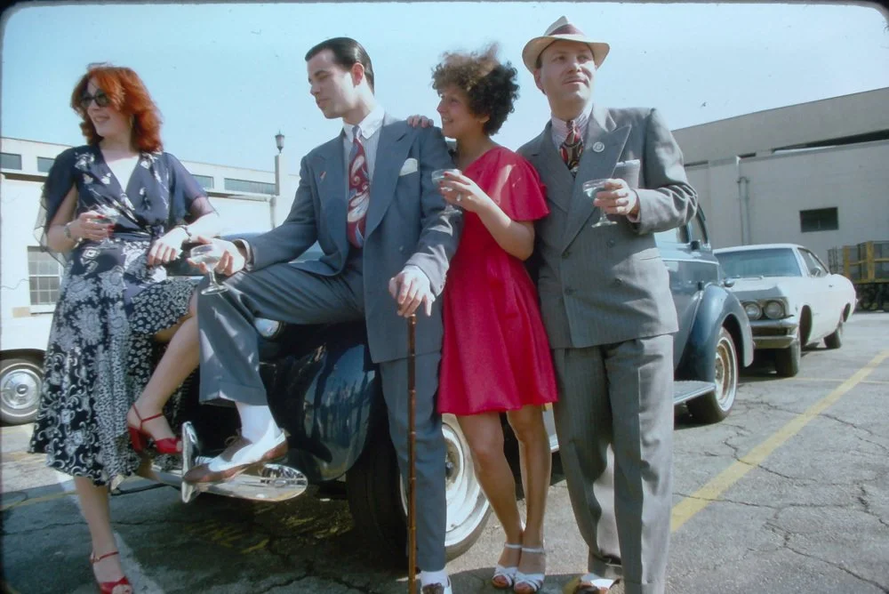 Four people dressed in vintage clothing standing and leaning against a classic car in a parking lot.