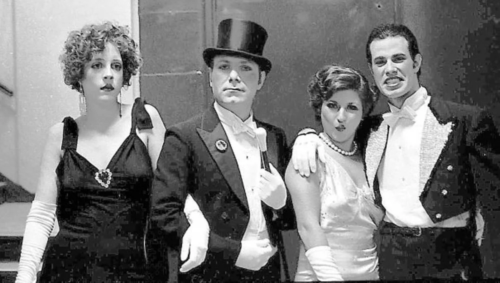 Four people in vintage formal attire, standing together at an event, with a woman on the left wearing a dress with a heart brooch, a man in a tuxedo with a top hat in the center, and a man and woman on the right wearing tuxedo and evening gown respec