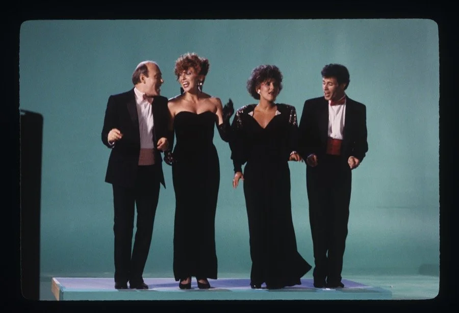 Four performers in elegant black tuxedos and evening gowns walking together on a stage with a plain teal background.