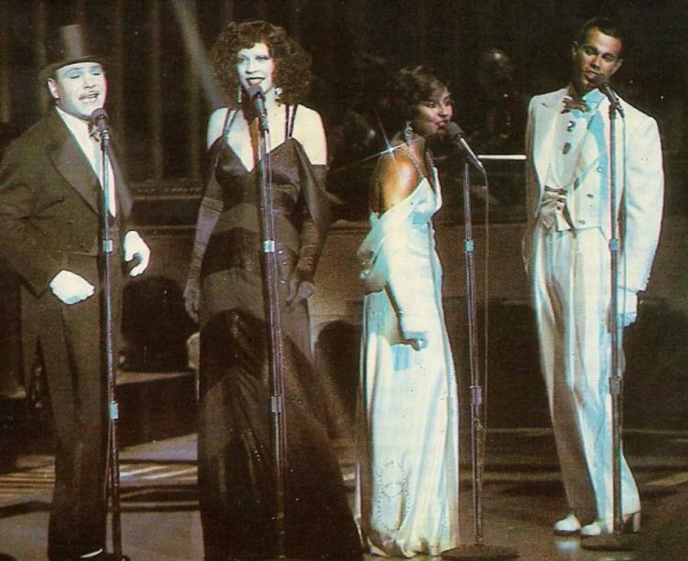 Four performers singing on stage with vintage microphone stands. The first performer on the left is dressed as a mime with white face paint, a top hat, and black suit. The second performer is a woman with curly hair in a black dress. The third perfor
