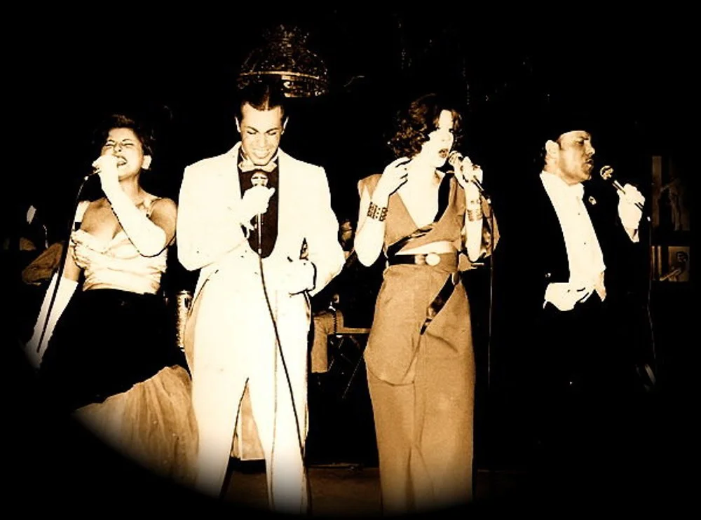 Four singers performing on stage, each holding a microphone, dressed in vintage formal attire, with a dark background and warm lighting.