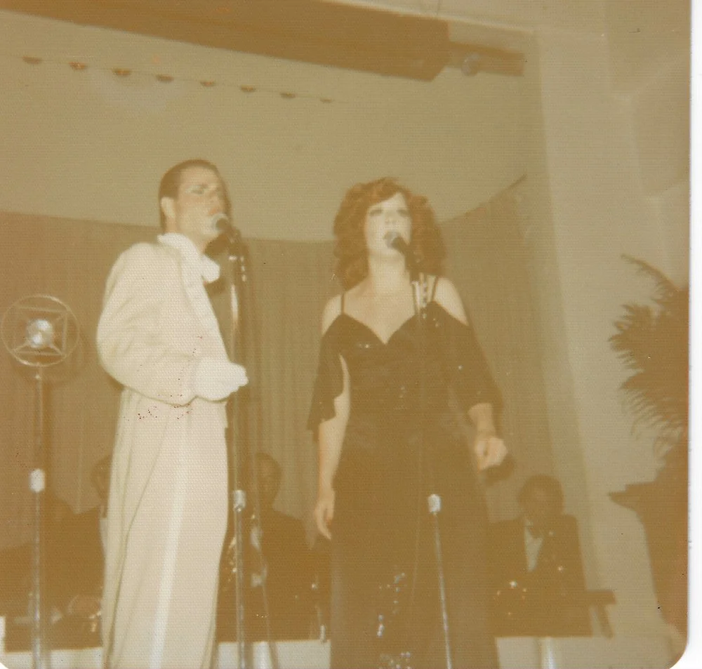 Two performers on stage, singing into microphones, dressed in vintage clothing with a backdrop and a potted plant visible in the background.