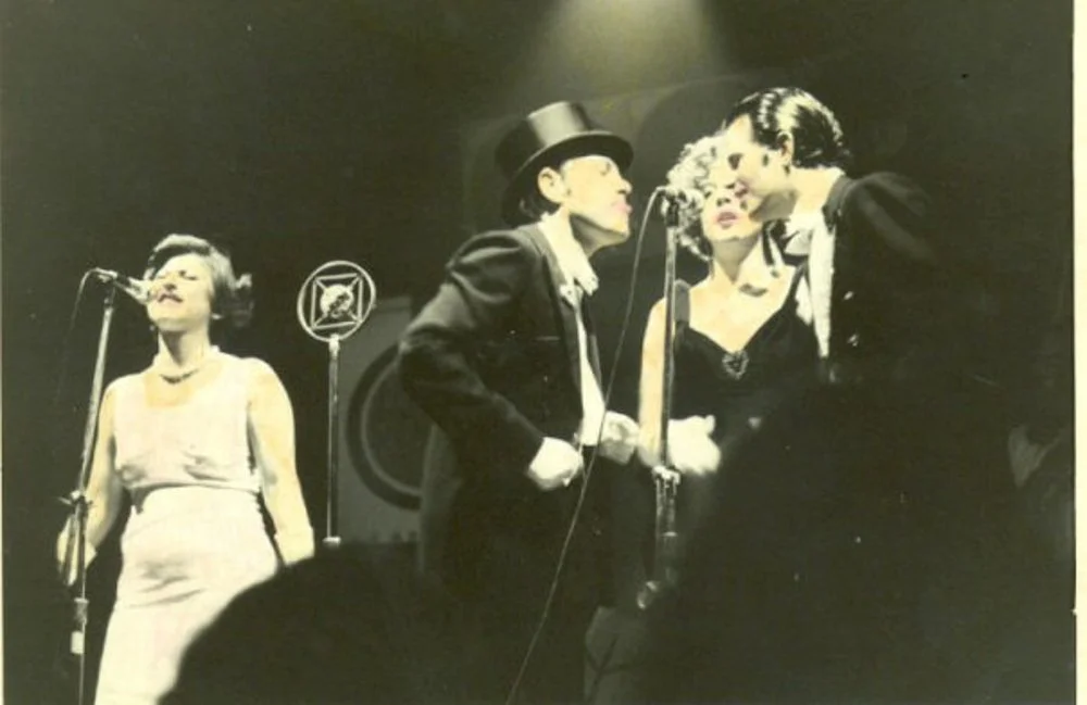 Four women on stage, one appears to be singing into a microphone, others are standing close together, in a performance setting with dark background.