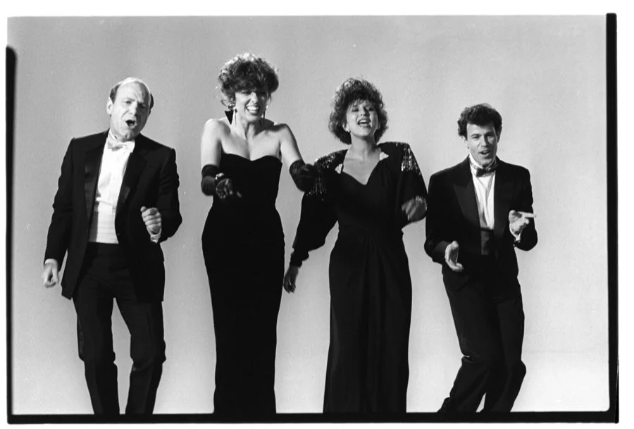 Four people in formal attire dancing and singing against a plain background.