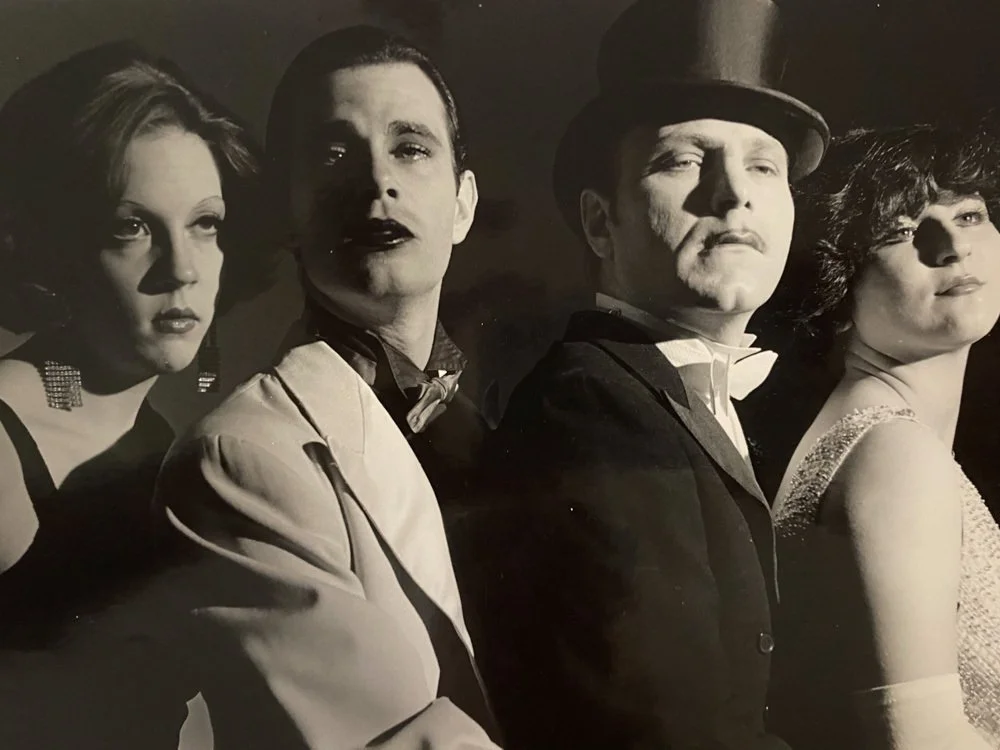 Black and white photo of four people, dressed in glamorous vintage-style clothing, posing with serious expressions. Two women and two men, one of the men wearing a top hat.