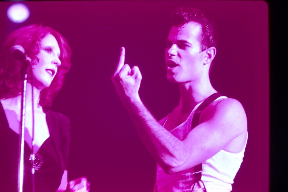 A man and woman having an intense conversation, with the man pointing his finger and the woman looking at him with a serious expression. The scene is lit with pinkish-purple lighting.