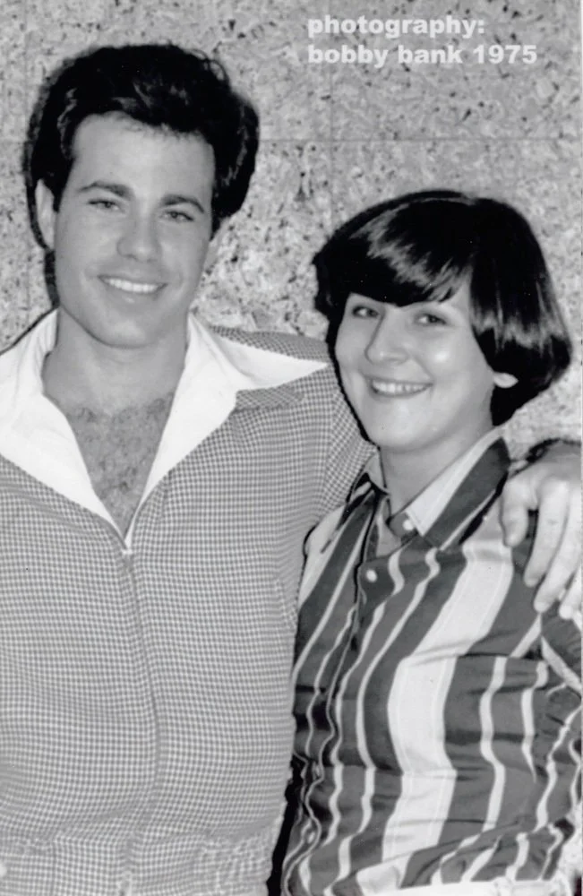 A black and white photo of a man and woman smiling and standing close together. The man has dark hair and is wearing a checkered shirt with a zip collar. The woman has a short haircut, is wearing a striped shirt, and has her arm around the man's shou