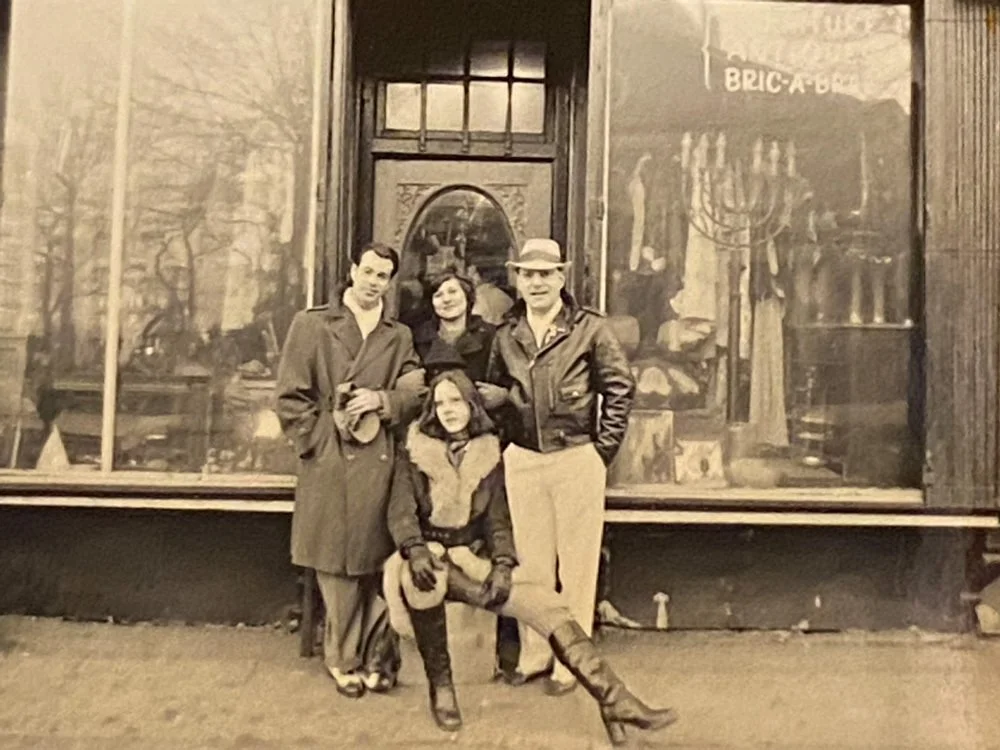Four people standing in front of a storefront window, which displays an interior scene with a menorah and mannequins. The group includes a woman, two men, and a girl holding a hat.