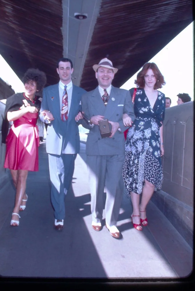 Four people walking under a bridge, smiling and holding hands. The woman on the left is wearing a pink dress and white sandals. The man next to her is dressed in a blue suit with a patterned tie. The man in the center is in a gray suit with a hat, ho