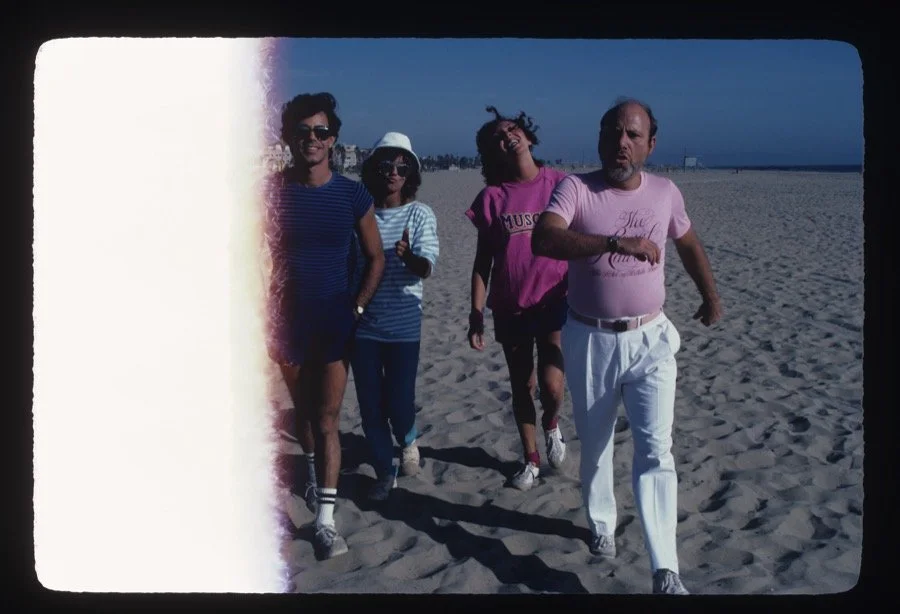Four people walking on a sandy beach near the ocean, dressed casually and enjoying the sunny day.