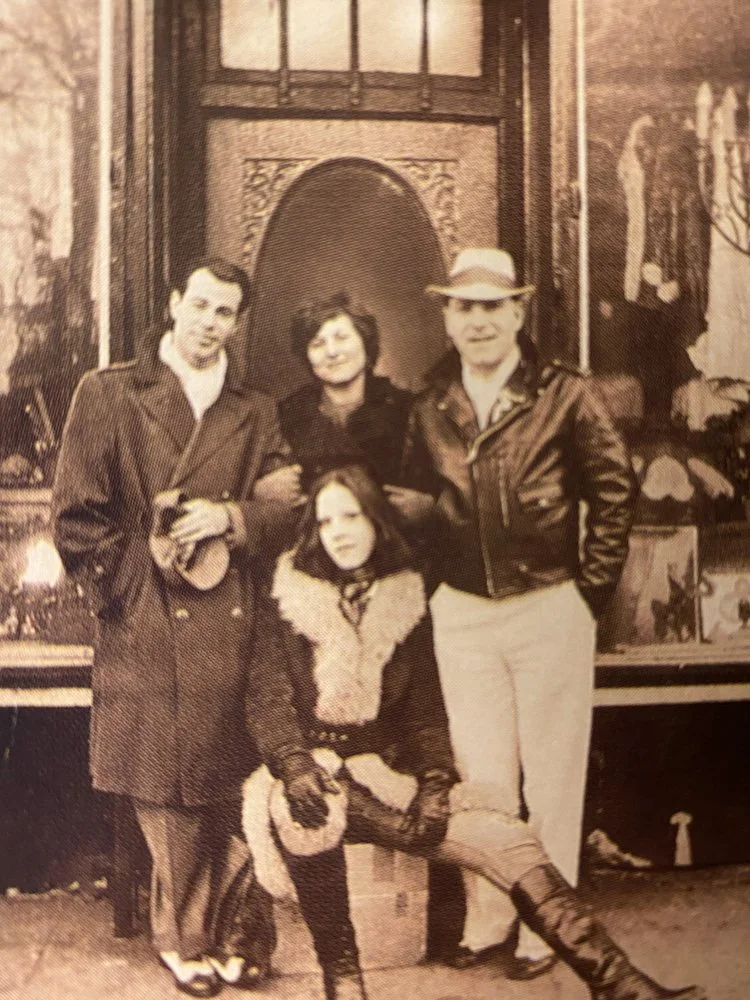 Four people posing in front of a decorative background, including two men and two women, with one woman seated on the ground, all dressed in vintage clothing.