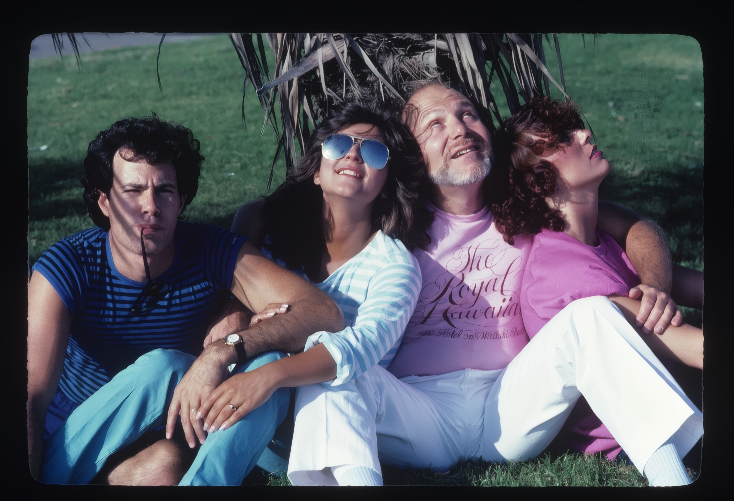 Four people lying on grass under a thatched roof, enjoying sunny weather.
