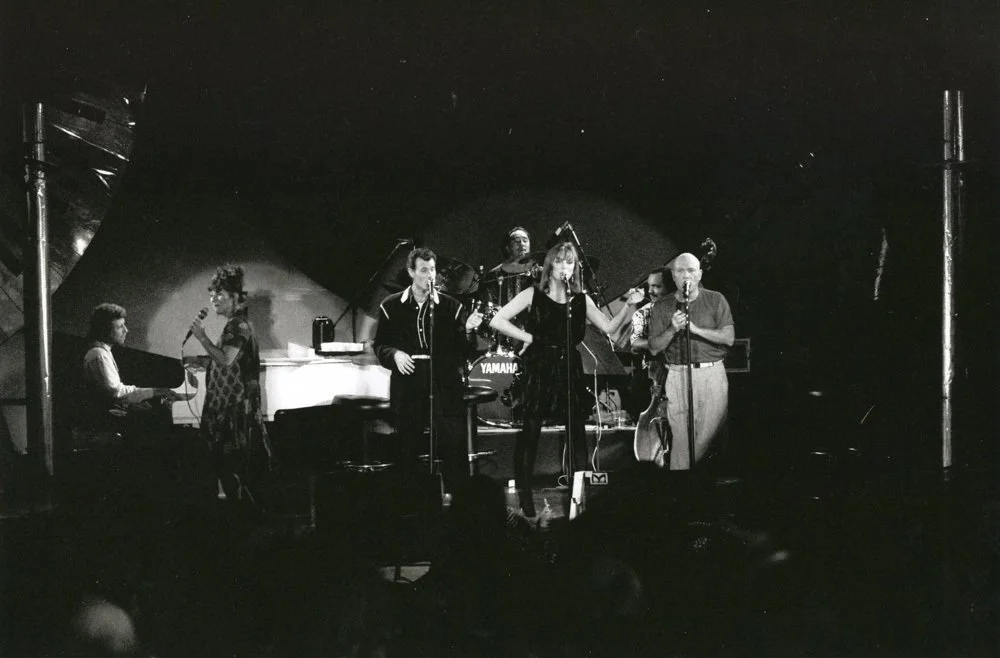 Black and white photo of a music band performing on stage, with a pianist, two vocalists, a drummer, and a saxophonist.