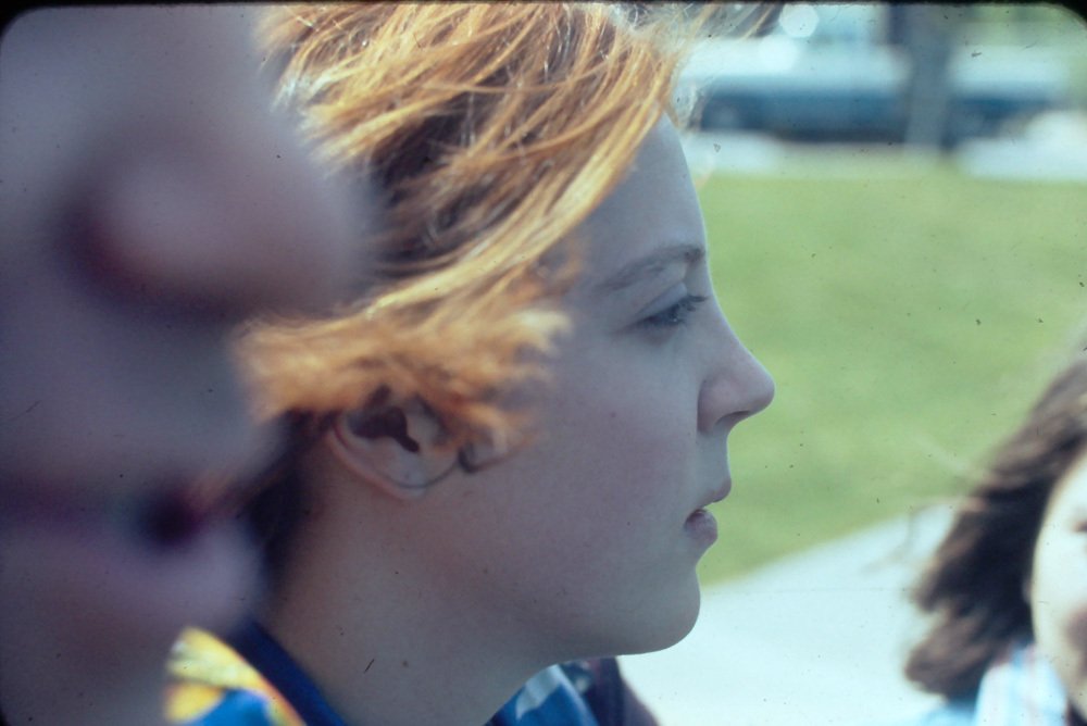 Profile of a young person with blond hair looking to the right, with some grass and blurred figures in the background.