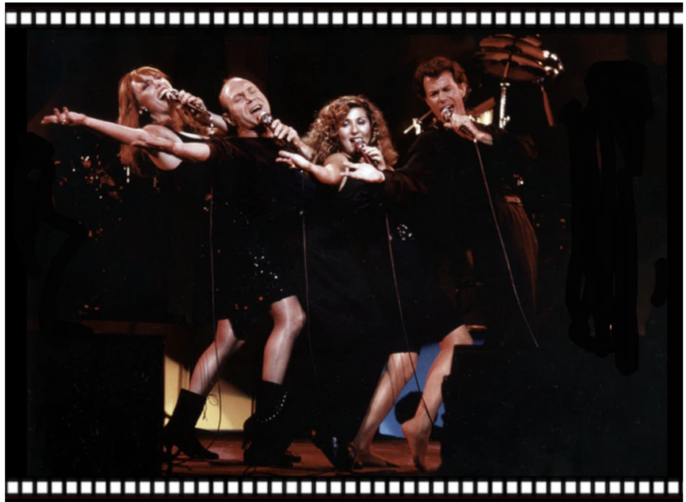 A group of four singers performing on stage, holding microphones, with their arms outstretched and singing passionately, dressed in black outfits, with a dark background and stage lighting.