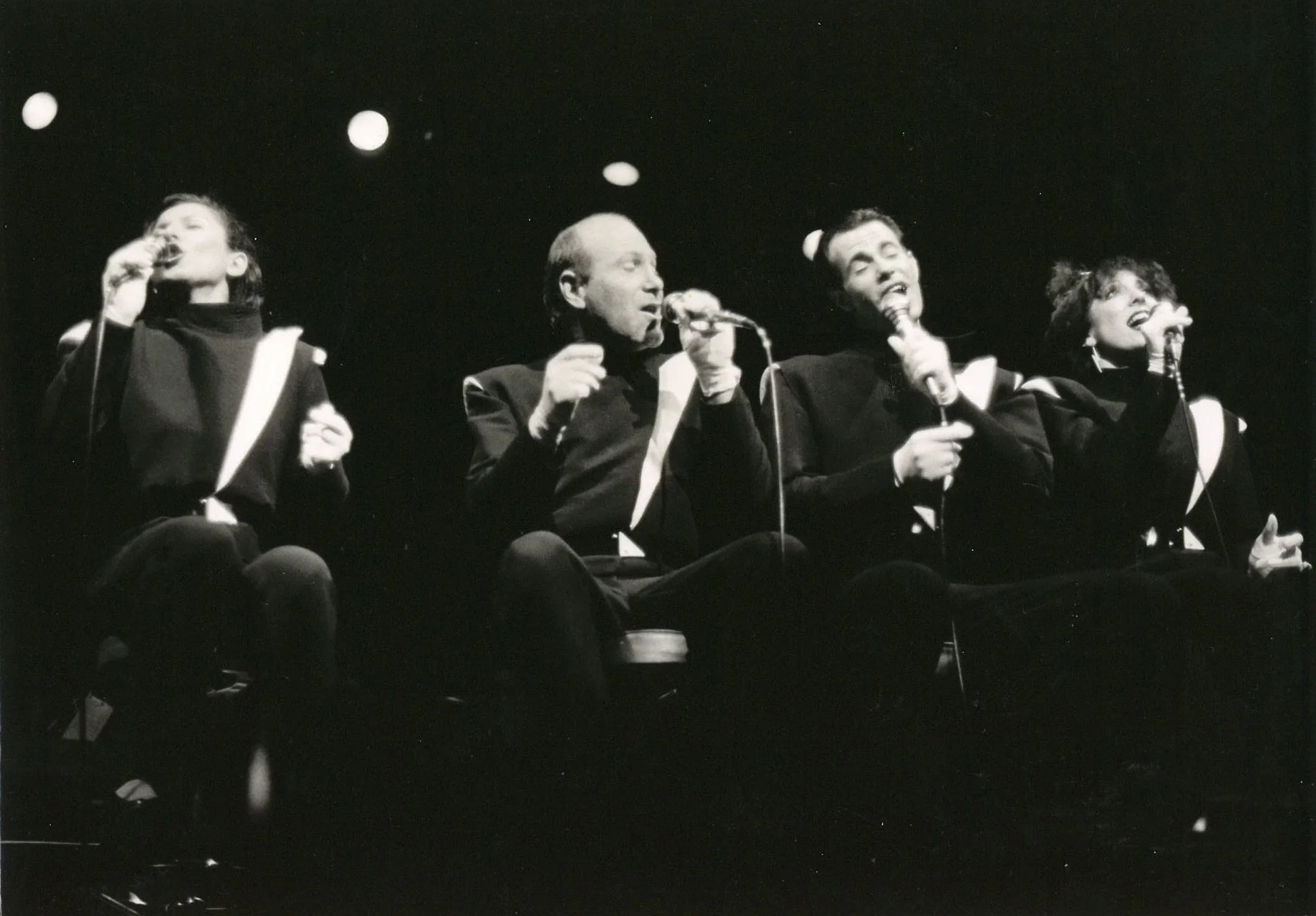 Black and white photo of four people sitting on a stage with microphones, singing or speaking, dressed in formal attire.