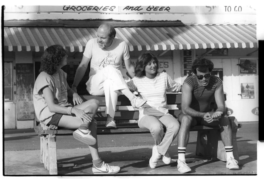 Four young people sitting on a bench in front of a store with a sign that reads 'Groceries and Beer.' They are engaged in conversation and appear to be enjoying a sunny day.