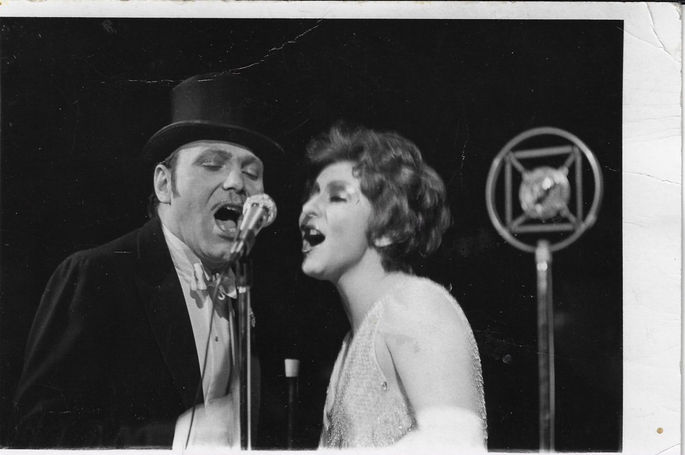 Black and white photo of a man and woman singing into a microphone, with a vintage studio microphone in front of them. The man is wearing a top hat and tuxedo, and the woman has short, curly hair and is wearing a light-colored dress.