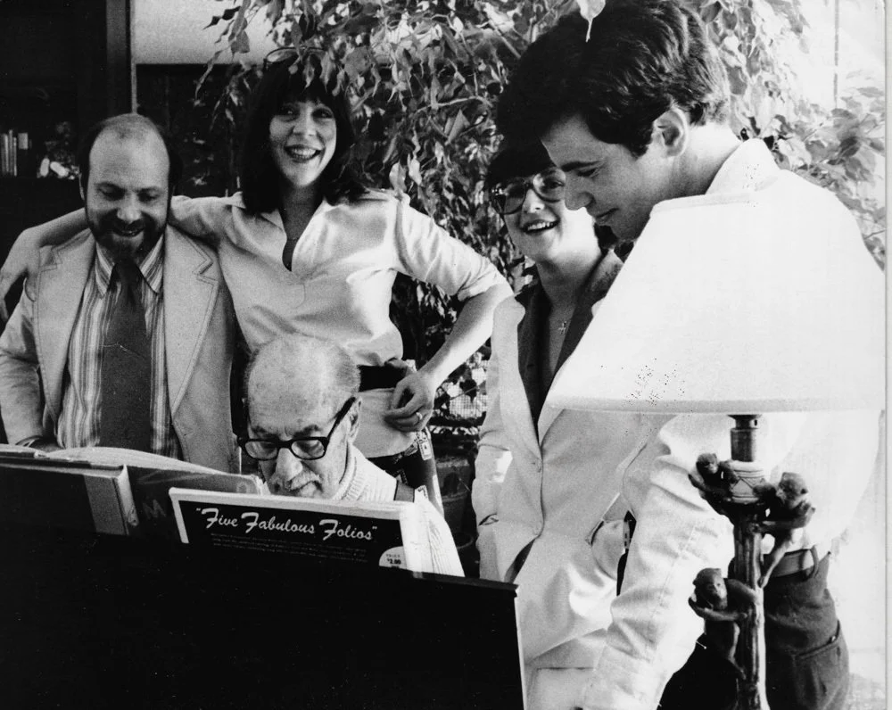 A group of five people gathered around a piano, with one man playing and the others watching and smiling. The scene appears to be in a cozy indoor setting, with a plant and a lamp nearby.