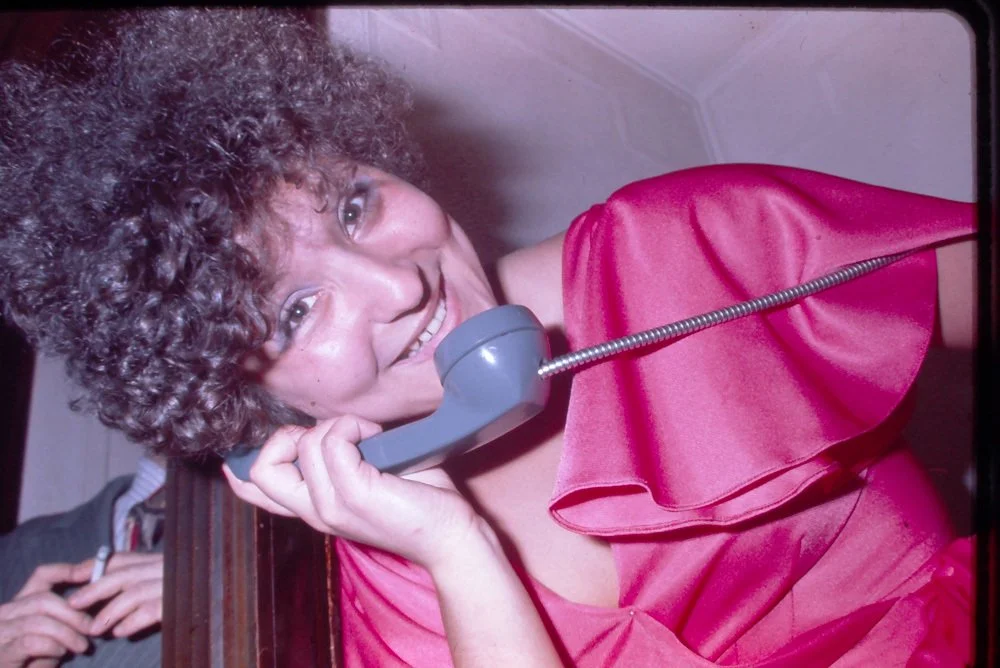 Woman with curly hair smiling and talking on a gray vintage telephone, wearing a shiny pink dress.