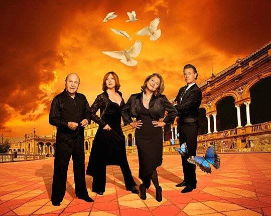 Four people standing on a patterned red stone pavement in front of an ancient colonnade with orange skies and white doves flying above, with a blue butterfly on one person's shoe.