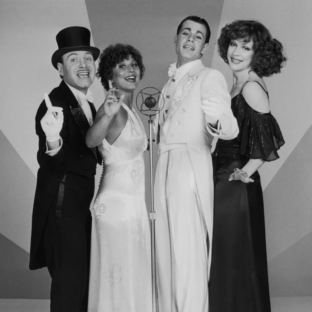 Black and white photo of five people in vintage formal attire, singing into a microphone, smiling and posing together.