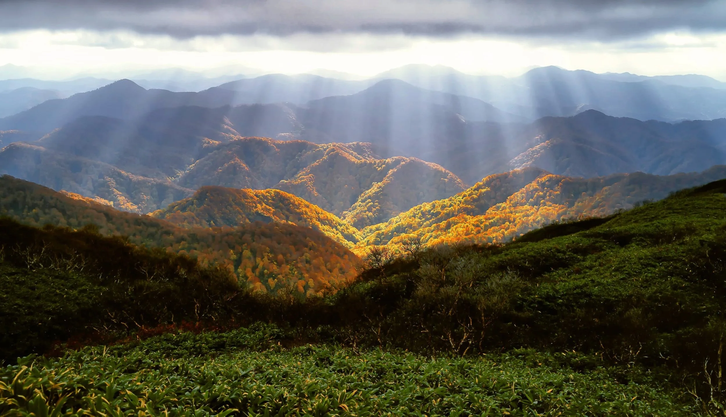 View of a mountain range with sunlight breaking through clouds, showcasing a mix of orange, green, and blue hues in the landscape.