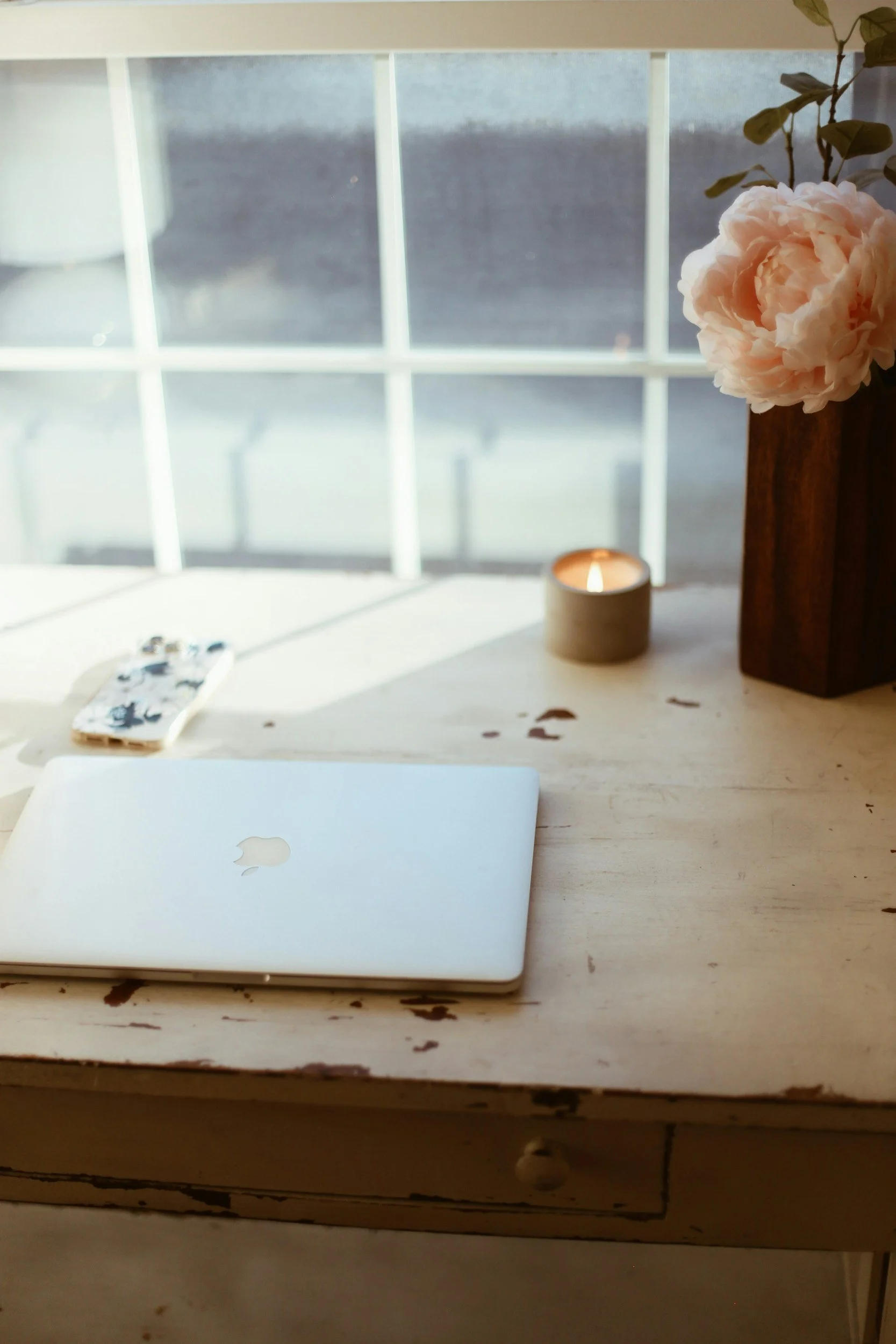 A white closed MacBook on a distressed wooden table with chipped paint, next to a small smartphone with a floral case, a burning candle in a cup, a vase with a pink flower, and a window in the background.