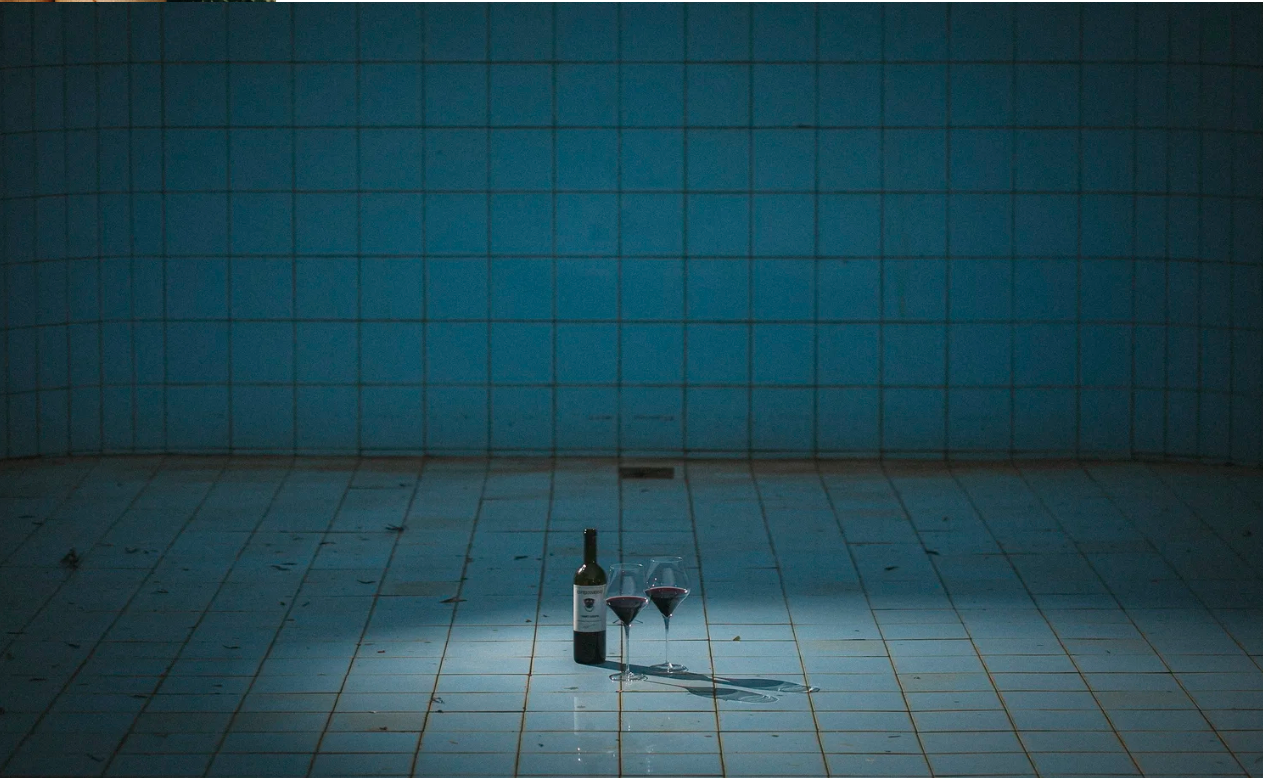 A bottle of red wine and two filled wine glasses on a tiled floor in front of a tiled wall, illuminated by a spotlight.