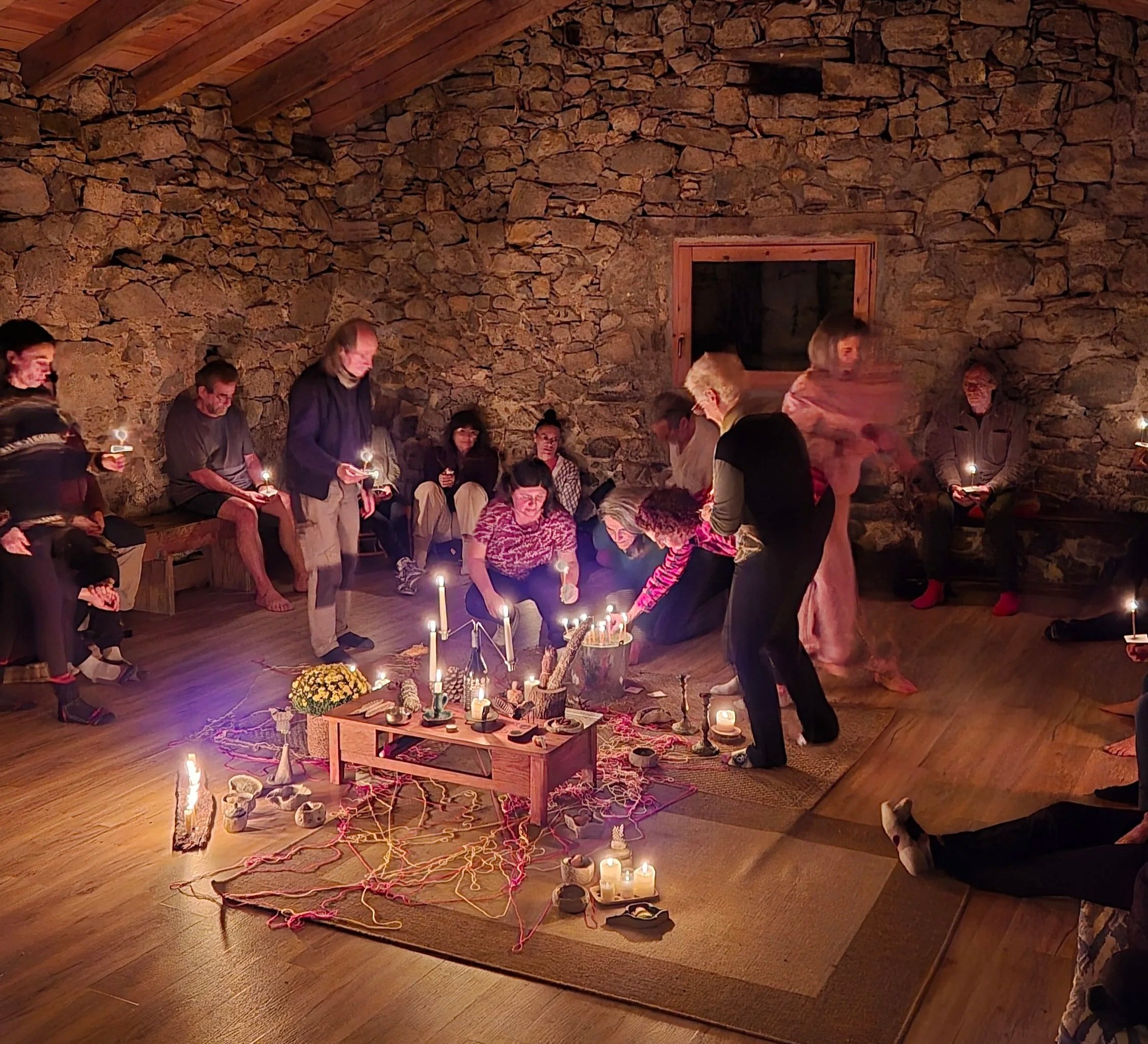 A group of people gathered around a table in a rustic room with stone walls and wooden ceiling beams. They are participating in a candle lighting ceremony, with various candles and decorative items arranged on the table and floor, along with flowers and candles.