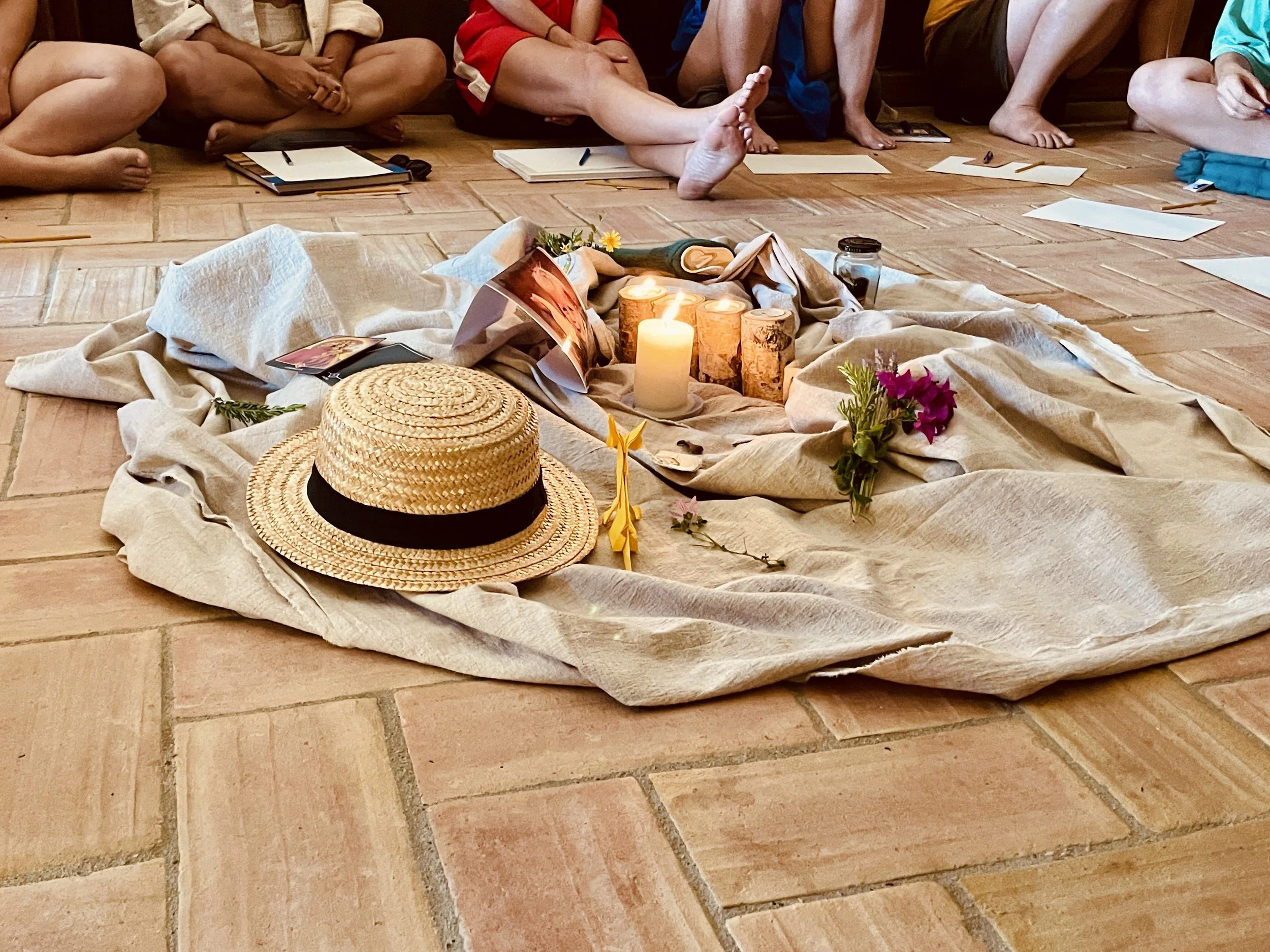 Group of people sitting cross-legged on the floor, surrounding a display of lit candles, flowers, and photographs, with notebooks and writing tools nearby.