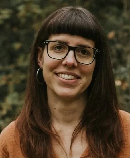 A woman with long brown hair, glasses, and hoop earrings outdoors with a blurred natural background.