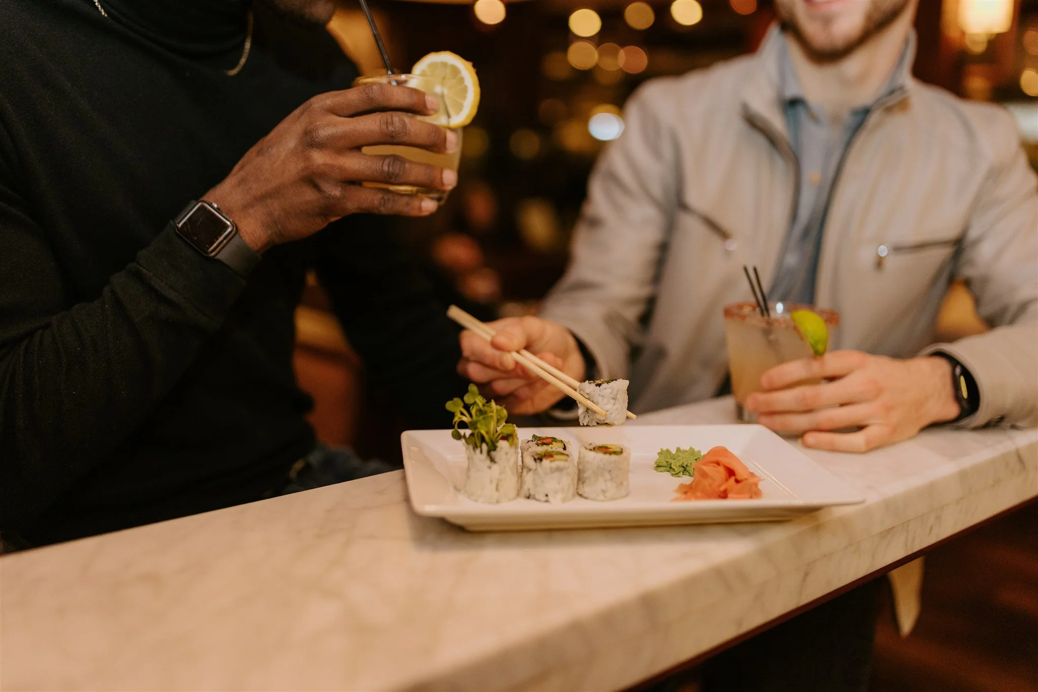 Two men having a drink and the white suited man picking a sushi with a chopstick