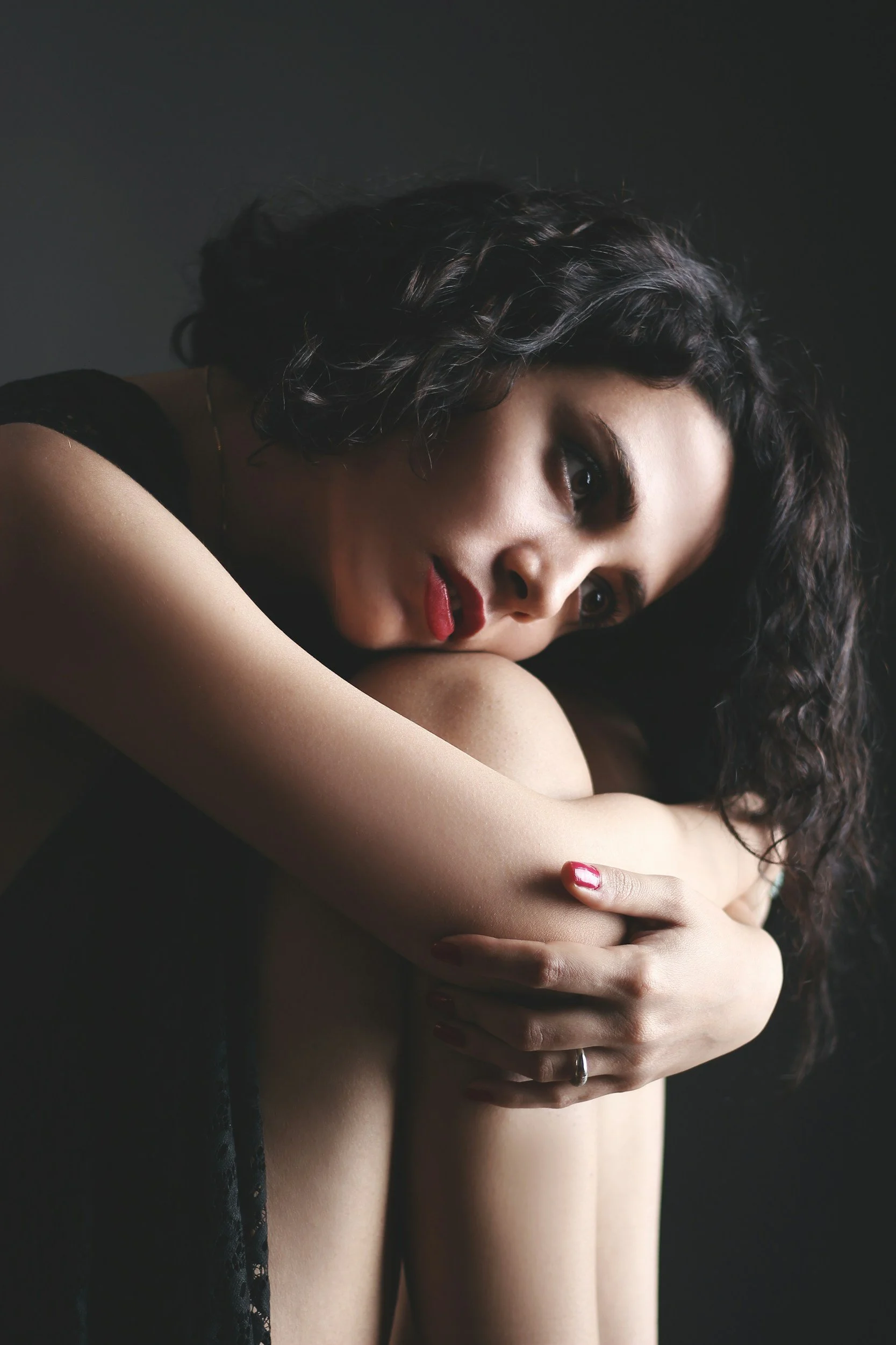 A woman with curly dark hair and red lipstick is holding her arm across her chest, partially covering her face, against a dark background.