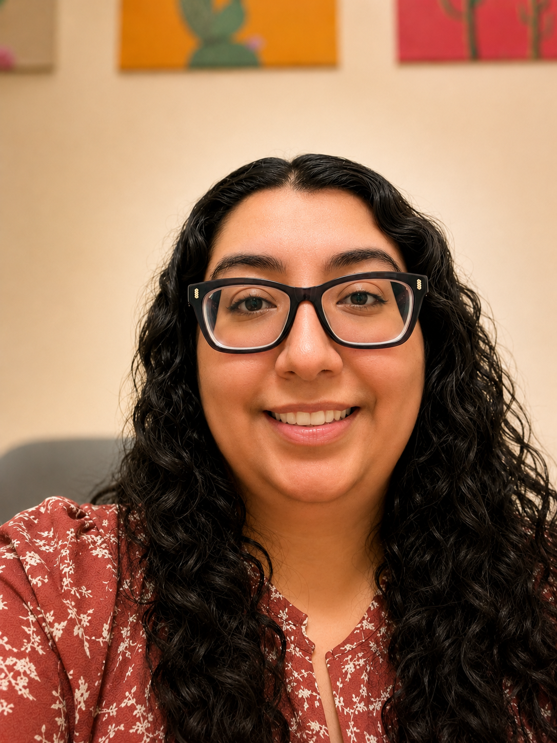 A smiling woman with long curly black hair and glasses, wearing a red patterned blouse, sitting in a room with colorful artwork on the wall behind her.