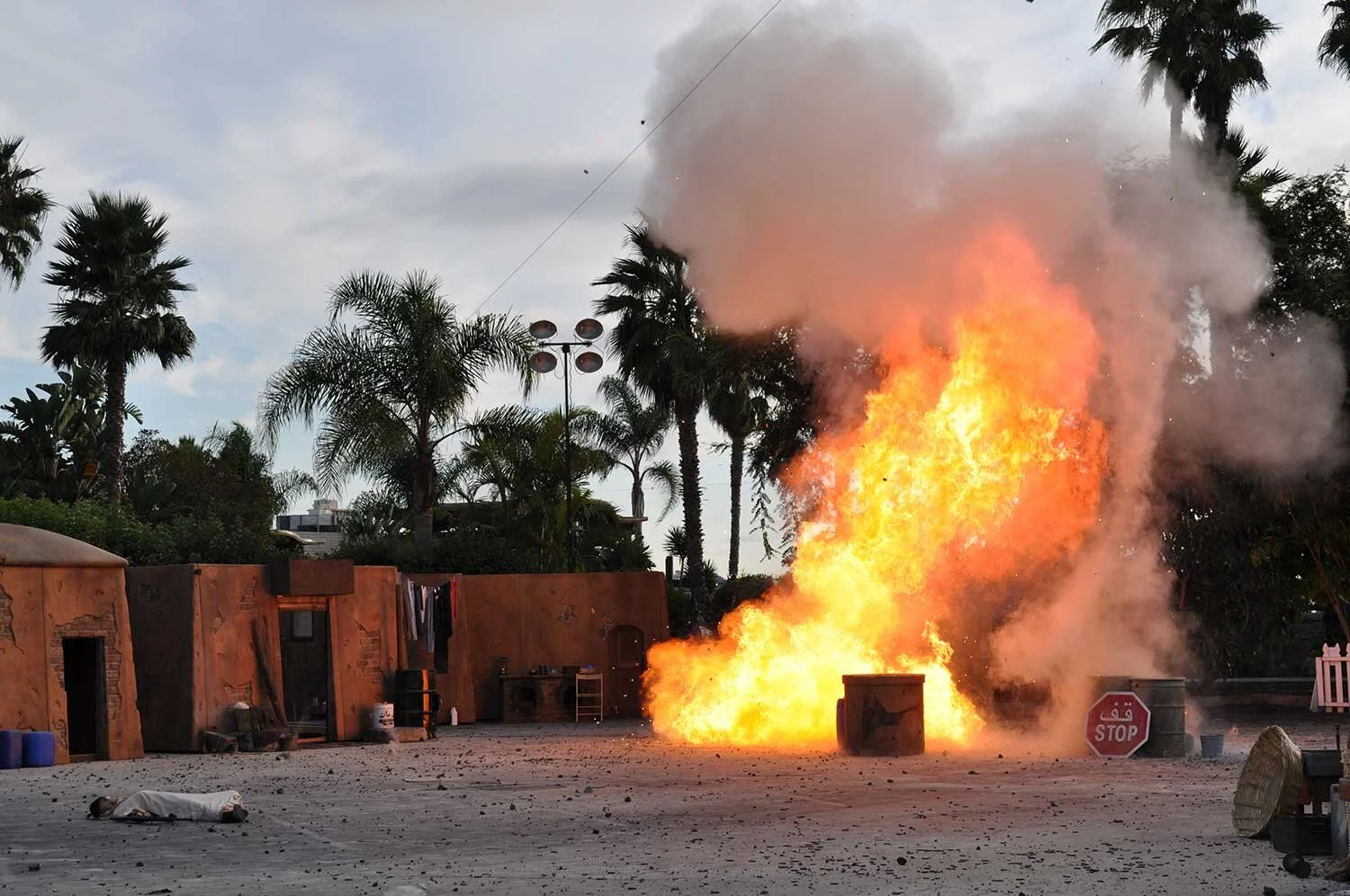 A large explosion with fire and smoke in an outdoor area surrounded by palm trees and buildings, with debris scattered on the ground and a person lying down nearby.