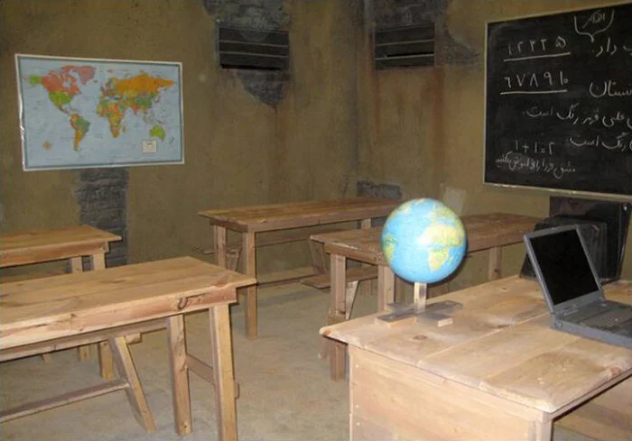 A classroom with five wooden desks, a blackboard with written text, a world globe on a desk, a laptop, and a world map on the wall.