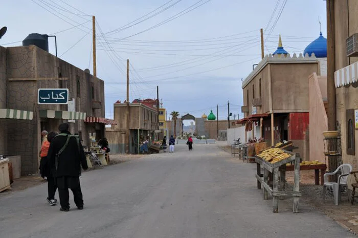 A quiet street in a Middle Eastern town with shops and pedestrians, some buildings have colorful domes, and there is an archway in the distance.