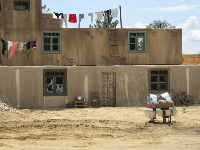 Two-story house with unfinished walls, green window frames, laundry hanging on a line, ladder on the upper level, and a cart with metal containers in front.