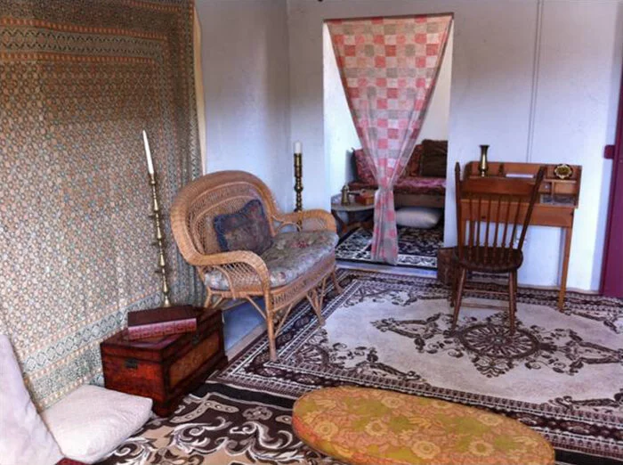 A vintage living room with a wicker loveseat, a wooden writing desk with a chair, a patterned area rug, and a canopy bed with pink checkered curtain.
