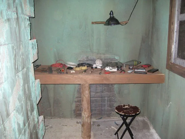 An unfinished green room with a wooden workbench cluttered with tools and objects, a small stool, and a hanging lamp.