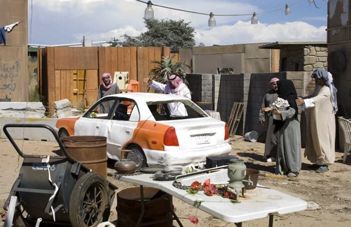 People wearing traditional Middle Eastern clothing surrounding a damaged car and various objects outdoors.